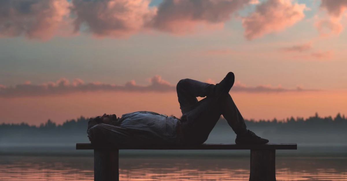 Man relaxing on a bench by a tranquil lake at sunrise, symbolizing rest, reflection, and mindful leadership.